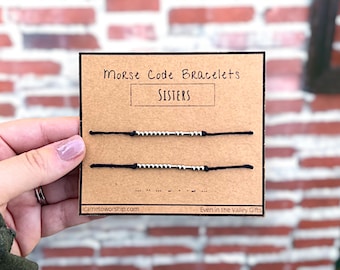 Puede incluir: Dos pulseras de cuerda negra con cuentas plateadas que deletrean la palabra "SISTERS" en código Morse. Las pulseras se muestran en una tarjeta marrón con el texto "Morse Code Bracelets" y "Even in the Valley Gifts".