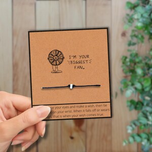 May include: A black and white wish bracelet with a card that says "I'M YOUR BIGGEST FAN." The card has a drawing of a fan. The text on the card says to make a wish and tie the bracelet on your wrist.