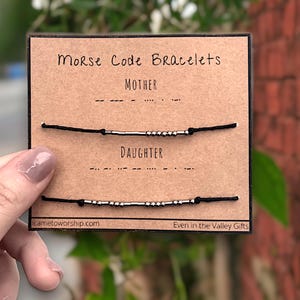 May include: A close-up of a card with the words "Morse Code Bracelets" printed on it. The card shows two black bracelets with silver beads that spell out "MOTHER" and "DAUGHTER" in Morse code. The card also includes the website "iametoworship.com" and the phrase "Even in the Valley Gifts".