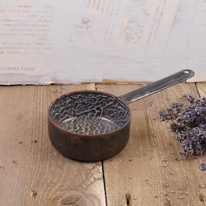 Op de afbeelding: Een vintage emaille pan met een zwarte handgreep en een gespikkelde wit-zwarte binnenkant. De pan staat op een houten oppervlak.