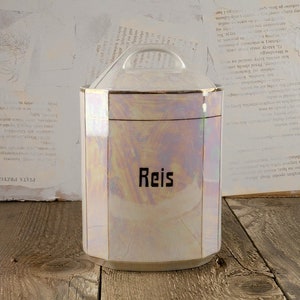 Op de afbeelding: Een witte keramische pot met een gouden rand en een deksel. De pot heeft een zeshoekige vorm en het woord "Reis" staat op de voorkant.