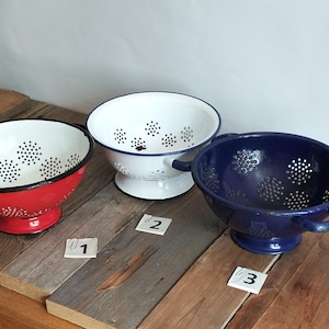 Puede incluir: Tres coladores de esmalte en rojo, blanco y azul, cada uno con un diseño perforado. Los coladores tienen asas y están sobre una superficie de madera. Los coladores están numerados 1, 2 y 3.