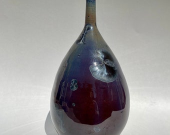 May include: A dark purple ceramic vase with a pearlescent glaze and white floral designs. The vase has a narrow neck and a rounded body.