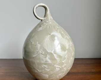 May include: A white ceramic vase with a unique, organic shape and a loop handle. The vase has a textured, crystalline glaze.