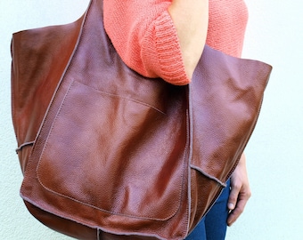 Puede incluir: Un bolso tote grande de cuero marrón con un bolsillo frontal. El bolso tiene una superficie texturizada y una parte superior ancha y abierta. El bolso se lleva sobre el hombro. La persona lleva un suéter color coral y vaqueros azules.
