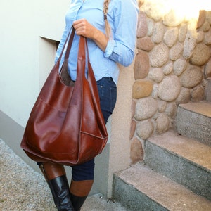 Puede incluir: Un bolso tote de cuero marr&oacute;n con un bolsillo grande en la parte delantera. El bolso lo lleva una persona que lleva una camisa de botones azul claro y pantalones vaqueros.