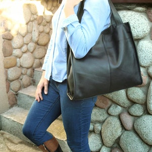 May include: A black leather tote bag with a rectangular shape and a shoulder strap. The bag is carried by a person wearing a light blue button-down shirt and blue jeans. The background features a stone wall.