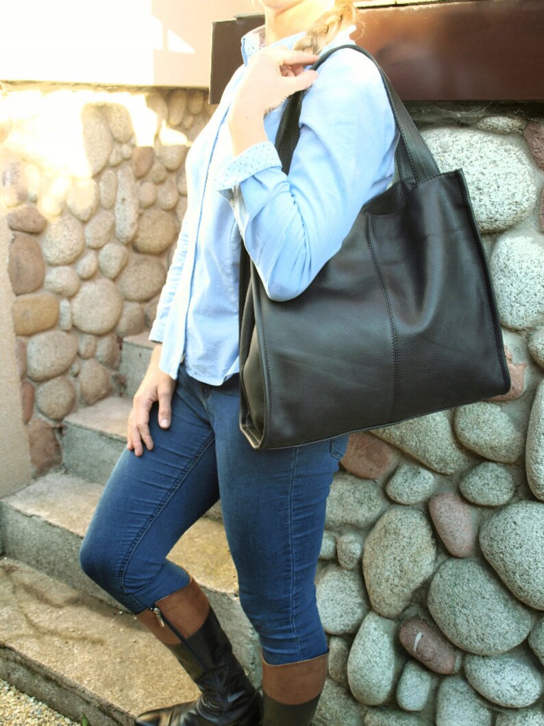 May include: A black leather tote bag with a rectangular shape and a shoulder strap. The bag is carried by a person wearing a light blue button-down shirt and blue jeans. The background features a stone wall.