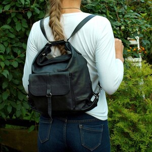 May include: Black leather backpack with adjustable straps and a front flap pocket. The bag has a rounded top and a structured design. The person is wearing a white long-sleeve shirt and blue jeans. The background features green foliage.