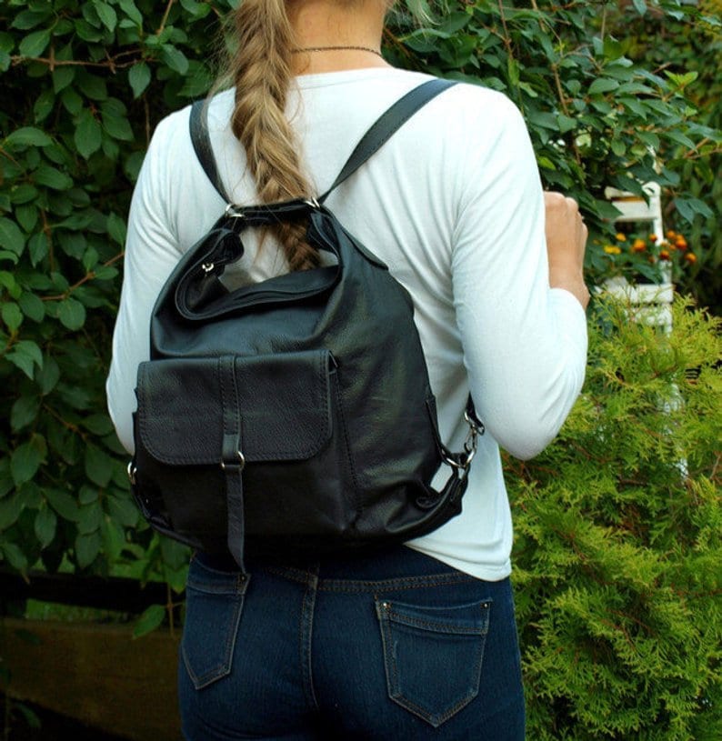 May include: Black leather backpack with adjustable straps and a front flap pocket. The bag has a rounded top and a structured design. The person is wearing a white long-sleeve shirt and blue jeans. The background features green foliage.