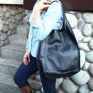May include: A woman wearing a light blue shirt and dark blue jeans is carrying a large black leather tote bag with a pocket on the front.
