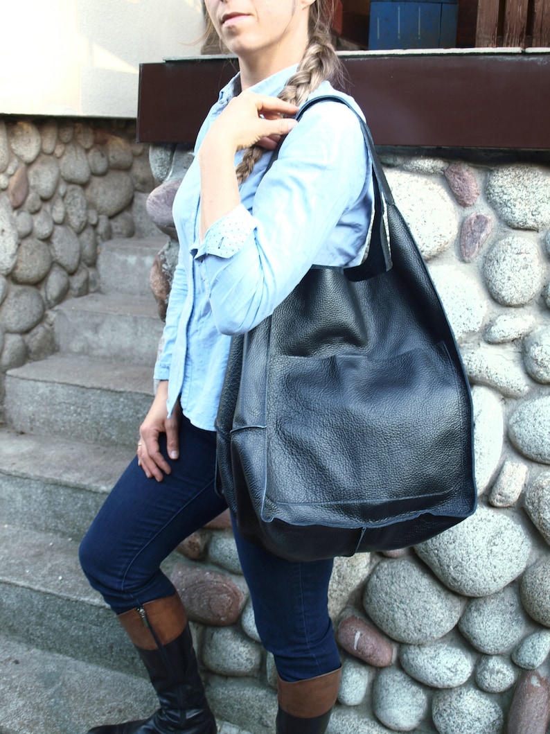 May include: A woman wearing a light blue shirt and dark blue jeans is carrying a large black leather tote bag with a pocket on the front.