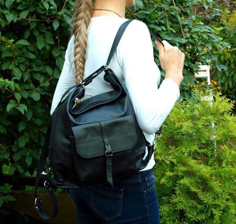 May include: Black leather backpack with a flap pocket and adjustable straps. The bag is worn over the shoulder. The person is wearing a white long-sleeve shirt and blue jeans. The background features green foliage.