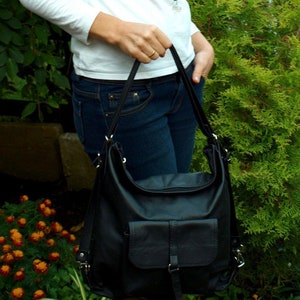 May include: Black leather shoulder bag with a front flap pocket and adjustable straps. The bag has a soft, slouchy design and is held by the straps. The background features green foliage and a person wearing a white shirt and blue jeans.