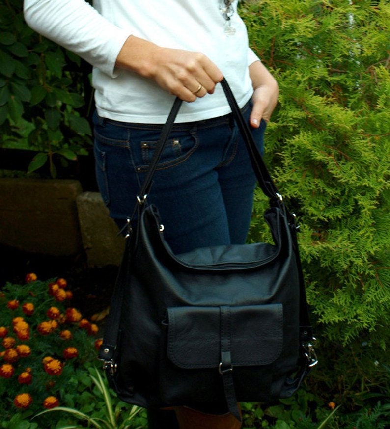 May include: Black leather shoulder bag with a front flap pocket and adjustable straps. The bag has a soft, slouchy design and is held by the straps. The background features green foliage and a person wearing a white shirt and blue jeans.