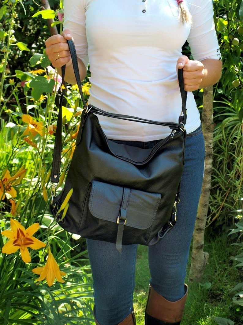May include: A black leather shoulder bag with a front pocket and adjustable strap. The bag has a flap closure with a buckle. The bag is held by a person wearing a white shirt and blue jeans.