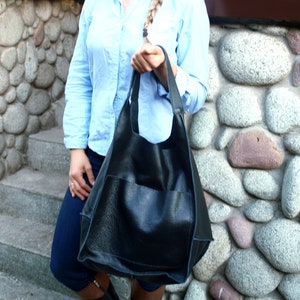 May include: A black leather tote bag with a large front pocket. The bag is being held by a person wearing a light blue shirt and dark blue trousers.