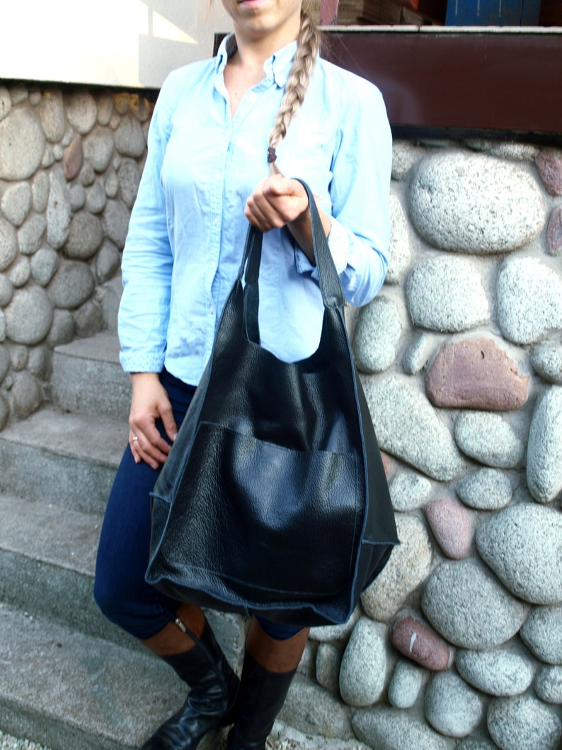 May include: A black leather tote bag with a large front pocket. The bag is being held by a person wearing a light blue shirt and dark blue trousers.