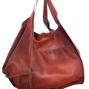 Puede incluir: Un bolso tote de cuero marr&oacute;n con un bolsillo &uacute;nico en la parte delantera. El bolso tiene una parte superior ancha y abierta y dos correas largas.
