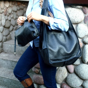 May include: A woman wearing a light blue button-down shirt and dark blue jeans carries two black leather bags. The larger bag is a slouchy tote bag with a long strap. The smaller bag is a rectangular crossbody bag.