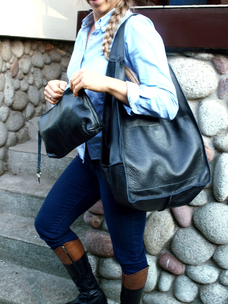 May include: A woman wearing a light blue button-down shirt and dark blue jeans carries two black leather bags. The larger bag is a slouchy tote bag with a long strap. The smaller bag is a rectangular crossbody bag.