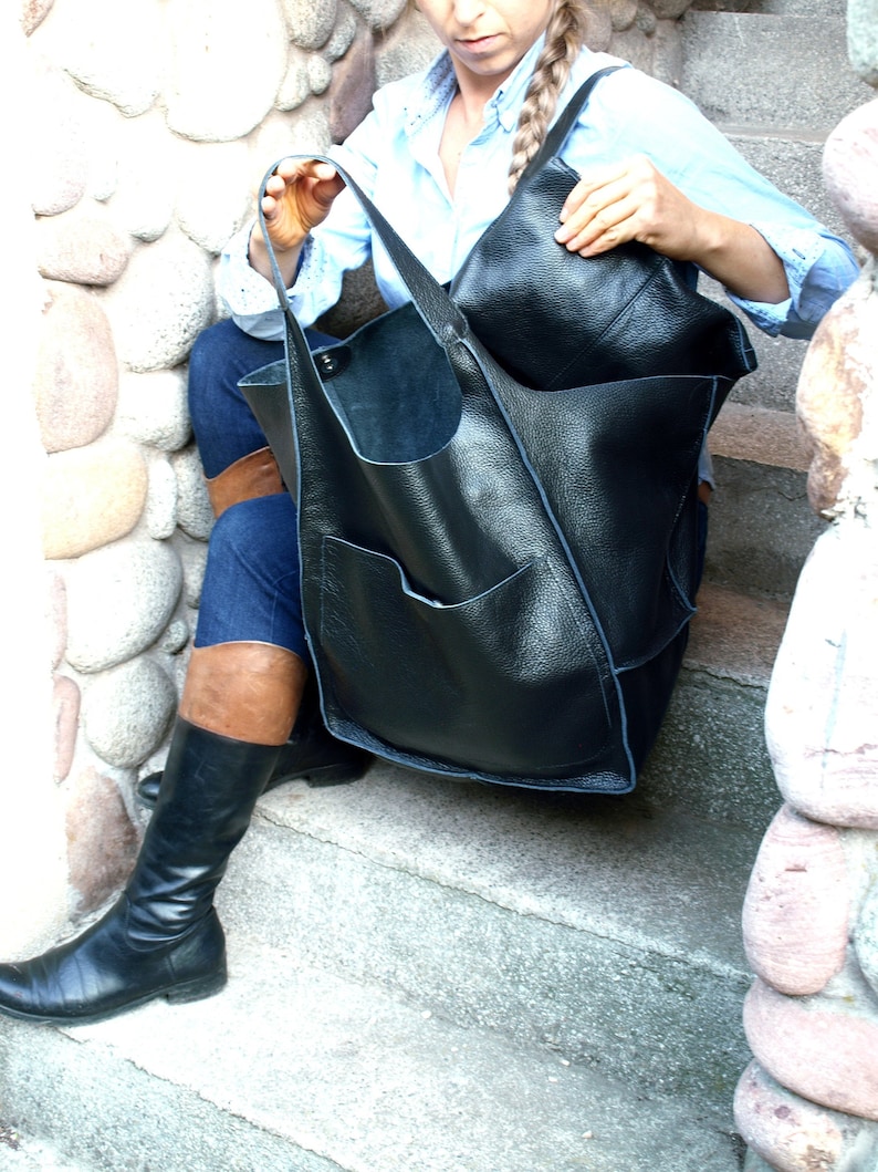 May include: A woman wearing blue jeans and black boots is holding a large black leather tote bag with a smaller black leather bag inside.