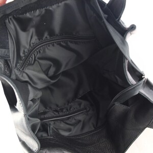 May include: Black leather tote bag with an open interior. The bag features a zippered pocket and a black fabric lining. The top of the bag has a velcro closure. The bag has two handles.
