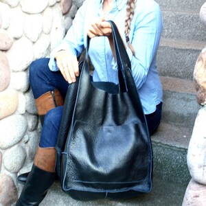 May include: A large black leather tote bag with a single pocket on the front. The bag has a wide opening and is made of textured leather.