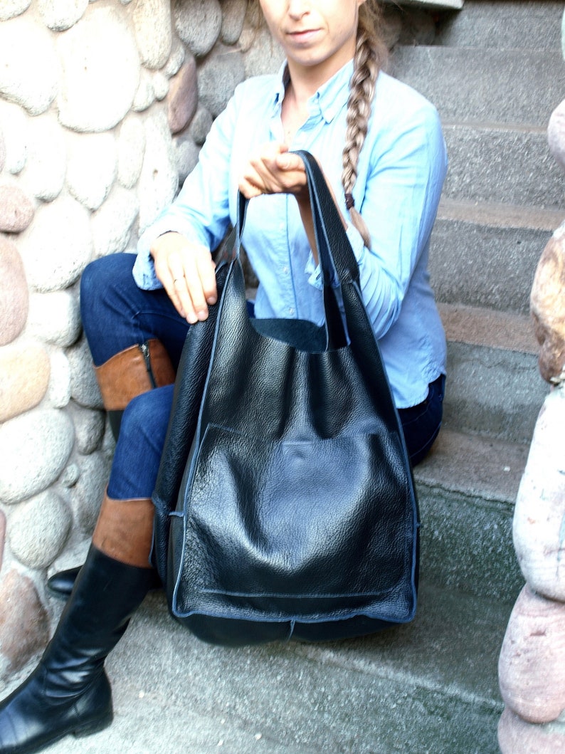 May include: A large black leather tote bag with a single pocket on the front. The bag has a wide opening and is made of textured leather.