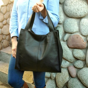 May include: A large, black leather tote bag with two handles. The bag is rectangular with a vertical seam down the center. The bag is being held by a person wearing a light blue button-down shirt and blue jeans.