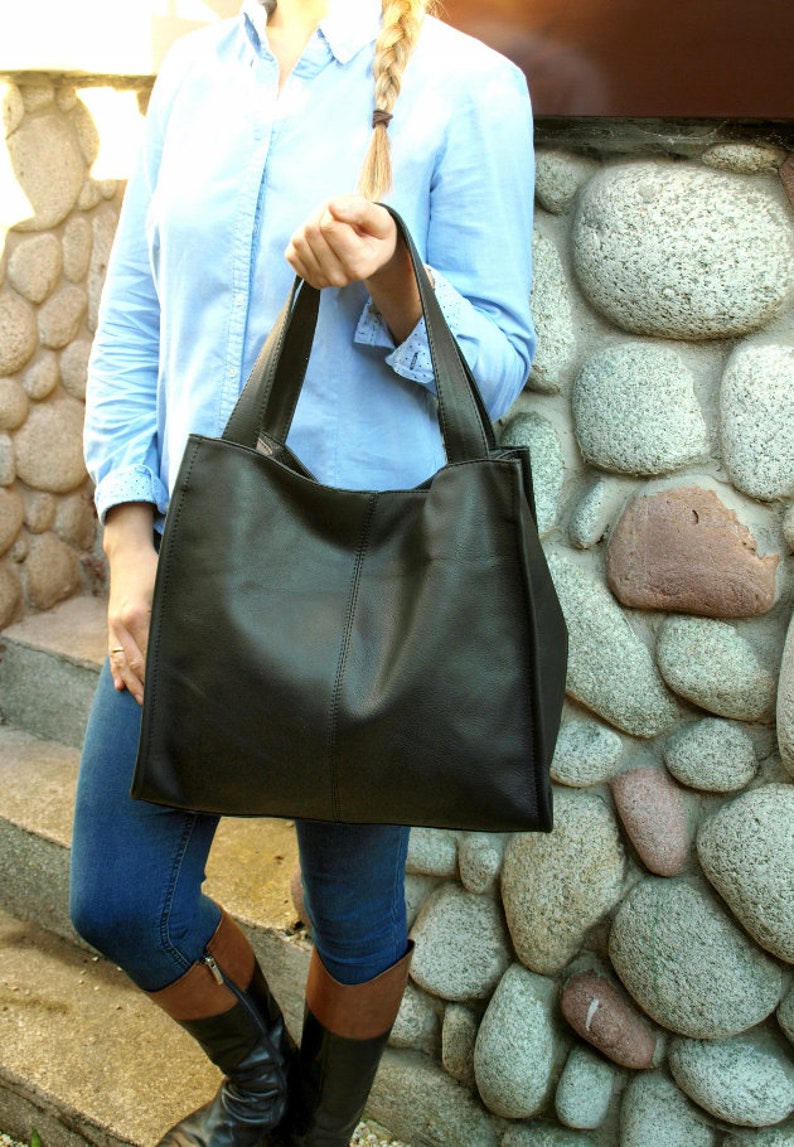 May include: A large, black leather tote bag with two handles. The bag is rectangular with a vertical seam down the center. The bag is being held by a person wearing a light blue button-down shirt and blue jeans.
