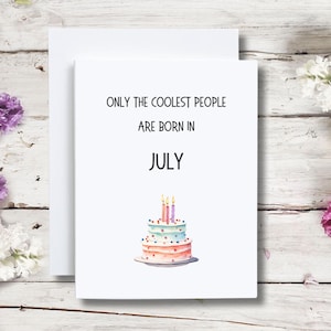 Op de afbeelding: Een witte kaart met zwarte tekst die "Only the coolest people are born in July" luidt. Onder de tekst staat een aquarel-illustratie van een drielaagse verjaardagstaart met roze, blauwe en witte frosting en brandende kaarsen.