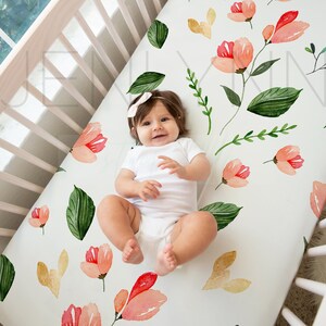 May include: A white crib sheet with a floral pattern featuring pink and green watercolor flowers and leaves. A baby is lying on the sheet.