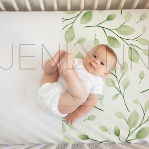 May include: A baby wearing a white onesie and a diaper lies on a white crib sheet with a green leafy pattern. The baby is looking up at the camera.