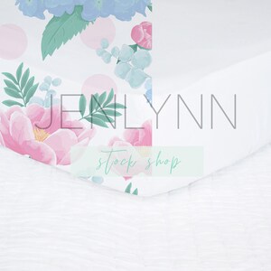 May include: White fitted crib sheet with a floral pattern featuring pink and blue flowers, green leaves, and white polka dots. The sheet has the text "JENLYNN stock shop" printed on it.