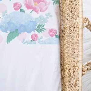 May include: A white crib sheet with a floral pattern featuring pink and blue flowers, green leaves, and pink polka dots. The sheet is partially tucked into a woven wicker basket.