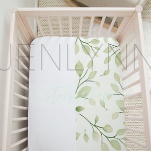 May include: A white crib with a white and green crib sheet featuring a leafy pattern. The crib sheet has the text "Jenlynn" printed on it.