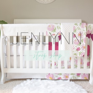 May include: White crib with a white and pink floral crib bedding set. The crib has a white bumper and a white sheet. The bedding set includes a pink and white floral blanket, a pink and white floral fitted sheet, and a pink and white floral crib skirt. The crib is in a nursery with a white rug and a green plant.