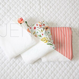 May include: Three white fabric swaddles with a floral print swaddle and a pink and white striped swaddle on top. The swaddles are arranged on a white quilted surface.