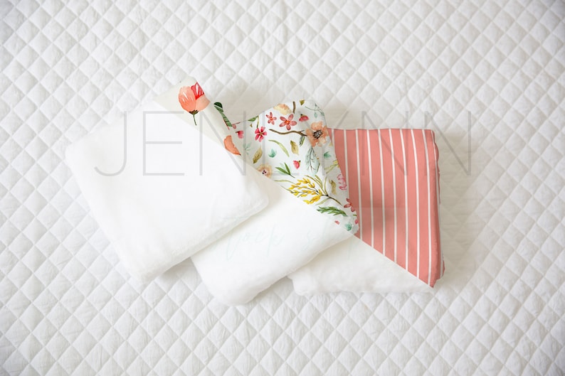 May include: Three white fabric swaddles with a floral print swaddle and a pink and white striped swaddle on top. The swaddles are arranged on a white quilted surface.
