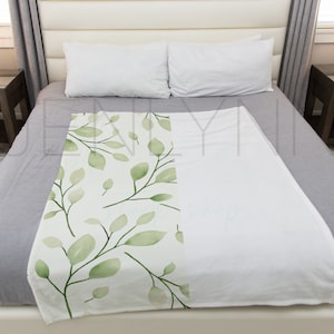 May include: A white throw blanket with a green leafy pattern on one side. The blanket is on a gray bed with white pillows.