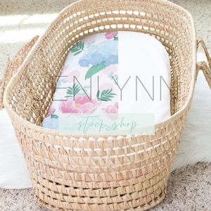 May include: A woven wicker bassinet with a floral patterned blanket. The blanket features pink and blue flowers with green leaves. The bassinet is on a white surface and a beige carpet. The text "NLYNN stock shop" is visible.