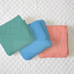 May include: Three folded fabric squares in teal, blue, and coral colors. The fabric squares are arranged in a stack on a white quilted surface.