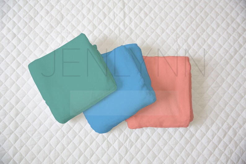 May include: Three folded fabric squares in teal, blue, and coral colors. The fabric squares are arranged in a stack on a white quilted surface.