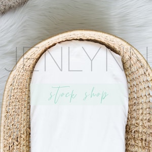 May include: Overhead shot of a woven wicker bassinet with a white sheet inside. The bassinet is set on a white faux fur rug and a neutral-toned carpet. The text "stock shop" is visible on the sheet.