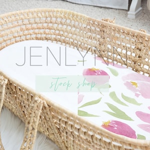 May include: A woven wicker bassinet with a white fitted sheet and a pink and green floral print. The sheet has a watercolor design with pink flowers and green leaves.