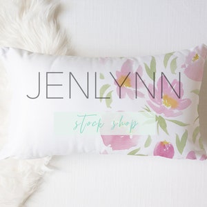 May include: Rectangular white pillow with a watercolor floral design in pink, yellow, and green. The pillow has the text "JENLYNN stock shop" printed on it. The pillow is on a white surface with a fluffy white faux fur.