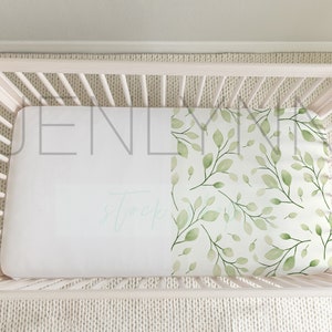 May include: White crib with a white and green fitted sheet with a leafy pattern. The sheet has the text "JENLYNN" printed on it.