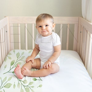 May include: A baby sitting in a white crib with a white fitted sheet and a green leaf patterned crib sheet. The baby is wearing a white onesie.
