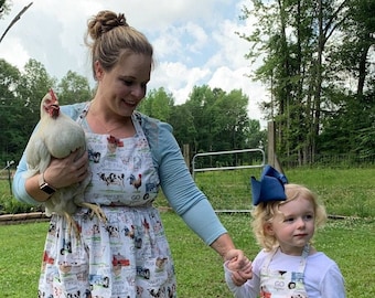 May include: A woman and a young girl wearing matching farm-themed aprons. The woman is holding a white chicken and the girl is holding a red basket with eggs. The aprons feature a white background with colorful farm animals and tractors.